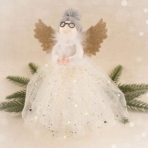 May include: A whimsical angel figurine with a gray bun, round glasses, and gold glitter wings. The angel wears a white dress with gold glitter accents and a pearl necklace. Decorative sprigs of evergreen are in the background.