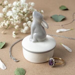 May include: A gray ceramic bunny figurine sits atop a white and gray trinket box. A gold ring with a purple gemstone and a silver necklace with a crescent moon charm are also visible. The background includes white flowers and green leaves.