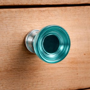 May include: A teal glass cabinet knob with a silver metal base, mounted on a light brown wooden surface. The knob is round with a dark centre and has a textured, rippled appearance. The knob's diameter is approximately 4 cm.