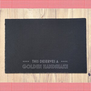 Op de afbeelding: Een rechthoekig zwart leisteen serveerbord met de tekst "THIS DESERVES A GOLDEN HANDSHAKE" in lichtgrijze letters. Het bord staat op een houten oppervlak, met een rood-witte geruite rand.
