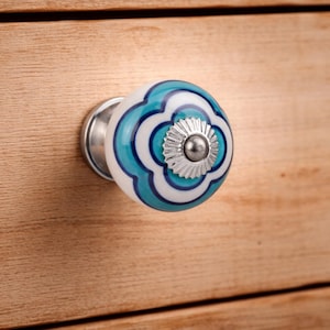 Op de afbeelding: Een ronde keramische meubelknop met een witte basis en concentrische, geschulpte patronen in turquoise en donkerblauw. Het midden van de knop is voorzien van een zilverkleurige metalen dop met een geribbelde rand en een kleine schroef.