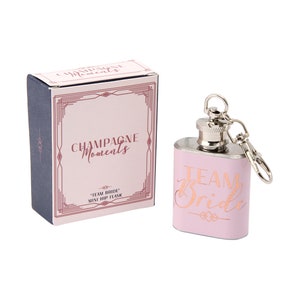 Op de afbeelding: Een roze metalen heupfles met een zilveren deksel en een zilveren ketting. De fles is gegraveerd met de woorden "Team Bride". De fles wordt geleverd in een doos met de woorden "Champagne Moments" en "Team Bride Mini Hip Flask" erop gedrukt.