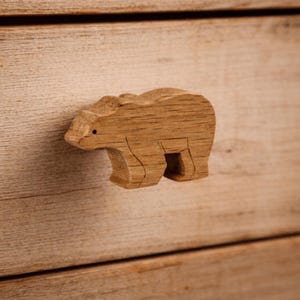 May include: A light brown wooden drawer knob carved into the shape of a standing bear. The bear figure features a visible wood grain pattern, a small dark dot for an eye, and incised lines defining the legs and body, attached to a wooden surface.