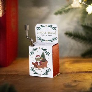 Pode incluir: Uma caixa de música branca com uma ilustração vermelha e verde de um urso usando um chapéu de Papai Noel puxando um trenó com uma árvore de Natal. A caixa de música tem as palavras "Jingle Bells Music Box" impressas na frente.