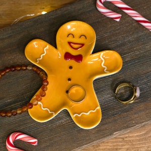 Pode incluir: Um prato de cerâmica amarelo em forma de homem de gengibre com um rosto sorridente e detalhes de bengala de caramelo vermelho e branco. O prato está sobre uma superfície de madeira com um colar de contas marrons e um anel de ouro.