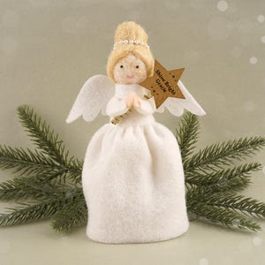 Op de afbeelding: Een handgemaakt engelenbeeldje met een witte jurk, vleugels en een halo. De engel houdt een stervormig label vast met de tekst "Shine Bright Gracie". De pop heeft blond haar en staat tegen een achtergrond van dennentakken.