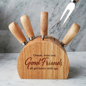Może przedstawiać: Drewniany zestaw noży do sera z czterema nożami i widelcem do sera, prezentowany w drewnianym bloku. Blok jest grawerowany słowami "Cheese, wine and Good Friends all get better with age."