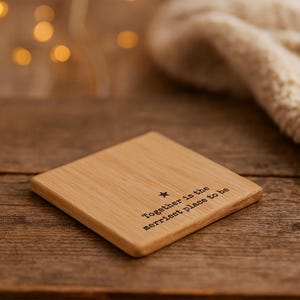 May include: Square wooden coaster with the text "Together is the merriest place to be" and a star symbol. The coaster is light brown and sits on a wooden surface. The background is blurred with warm tones.
