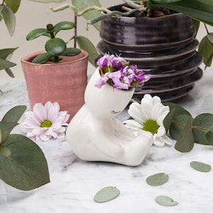 May include: White ceramic planter shaped like a cat with a small bouquet of purple flowers inside. The planter is sitting on a white marble surface with green leaves scattered around it.