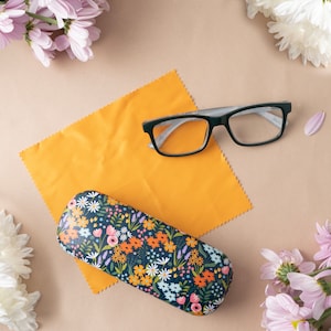 May include: Rectangular eyeglasses with a black and grey frame rest on a yellow microfiber cloth. A floral patterned glasses case in navy blue, orange, and white sits below. Pink and white flowers surround the items.