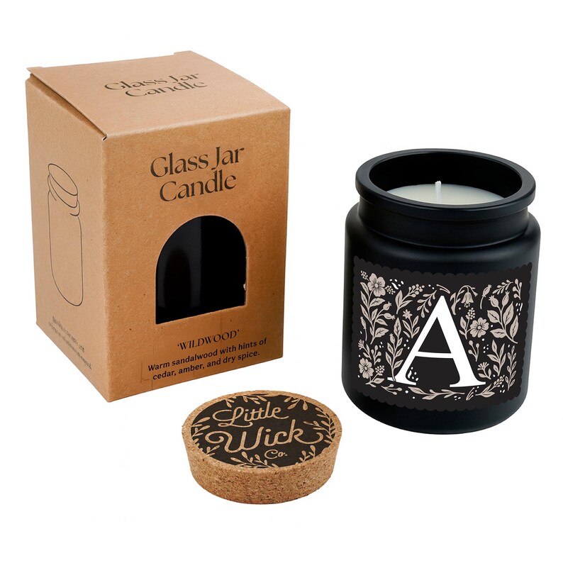 Puede incluir: Una vela en tarro de cristal negro con una mecha blanca, una tapa de corcho y una caja de cart&oacute;n. La vela tiene una "A" blanca sobre una etiqueta floral. La caja y la etiqueta de la vela dicen "Glass Jar Candle" y "Wildwood". La tapa de corcho dice "Little Wick Co."