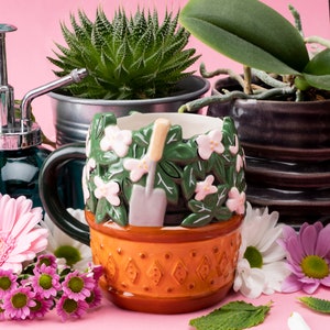 May include: A ceramic mug shaped like a flower pot with pink flowers and green leaves. The mug has a brown handle and a brown base with a diamond pattern. The mug is sitting on a pink surface with other plants and flowers.