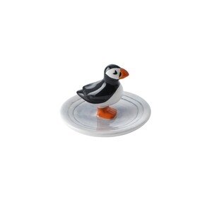 May include: A ceramic puffin figurine with black, white, and orange coloring, perched on a small, round, gray plate. The bird has a detailed beak and stands upright. The plate has a simple, minimalist design.