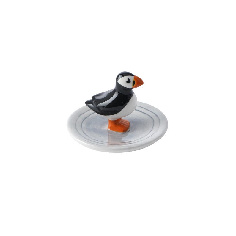 May include: A ceramic puffin figurine with black, white, and orange coloring, perched on a small, round, gray plate. The bird has a detailed beak and stands upright. The plate has a simple, minimalist design.