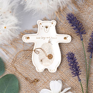May include: White ceramic bear-shaped trinket dish with gold accents. The dish has the words "bear hugs and kissus" written on it. The dish is ideal for holding jewellery, rings, or other small items.