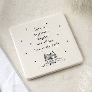 Op de afbeelding: Witte keramische onderzetter met zwarte tekst en een eenvoudige huisillustratie. De tekst luidt "home is happiness, laughter, and all the love in the world."
