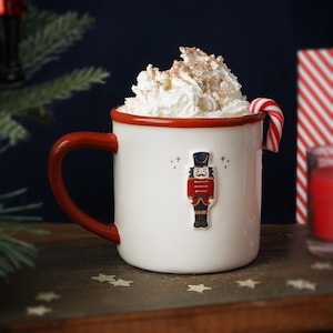 Pode incluir: Caneca de cerâmica branca com borda e alça vermelhas. A caneca apresenta um design festivo de quebra-nozes e está cheia de uma bebida quente coberta com chantilly e um bastão de caramelo.