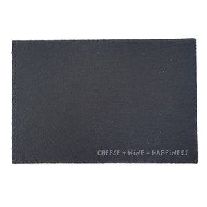 Puede incluir: Tabla de queso rectangular de pizarra negra con bordes rugosos. La frase "CHEESE + WINE = HAPPINESS" está impresa en gris claro en la esquina inferior derecha. La tabla está aislada sobre un fondo blanco.