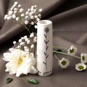 Könnte beinhalten: Weiße Keramikvase mit einer schwarzen Linienzeichnung einer Blume und dem Text "Let me grow". Die Vase steht auf einer braunen Stoffoberfläche mit weißen Blumen.