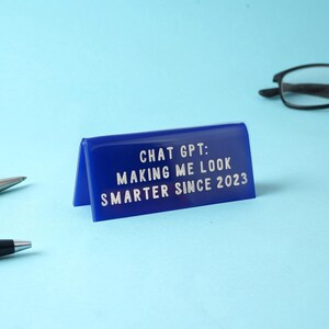 May include: A blue desk sign with the text "CHAT GPT: MAKING ME LOOK SMARTER SINCE 2023" in white lettering. The sign is angled, with a light blue background. Pens and glasses are visible in the background.