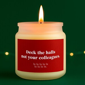 May include: A lit, cream-coloured candle in a frosted glass jar. A red label displays the text "Deck the halls not your colleagues. la la la la la la la la la." The candle has a bright yellow flame against a green background.