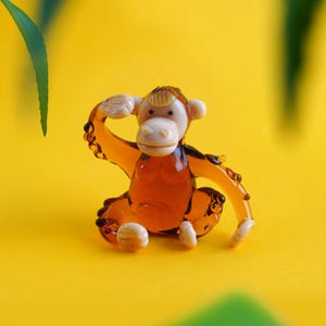 以下が含まれることがあります： 琥珀色の小さなガラス製の猿の置物。顔と手は薄いベージュ色。猿は片手を頭に当ててポーズをとっており、体には小さな透明なガラスのアクセントが施されています。明るい黄色の表面に置かれています。