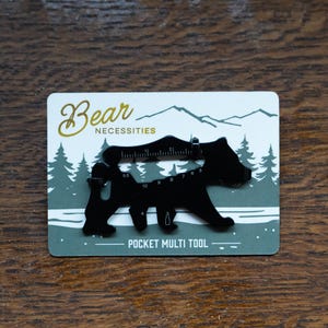 Può includere: Un multiutensile tascabile nero a forma di orso, con un righello e varie funzioni. L'utensile è su una carta con il testo "Bear Necessities" e "Pocket Multi Tool", su uno sfondo di alberi e montagne.