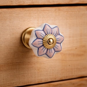 May include: A flower-shaped cabinet knob featuring white ceramic petals with soft pink centers and blue outlines. The center of the flower is a circular gold-toned metal piece with a ridged edge, and the base is a smooth gold-toned metal cylinder.