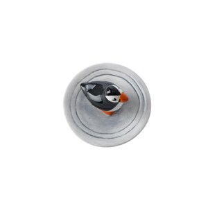 May include: A ceramic puffin figurine with a black and white body, orange beak, and white eye markings. The bird is perched on a round, gray, textured base with concentric circle patterns. The figurine is a decorative item.