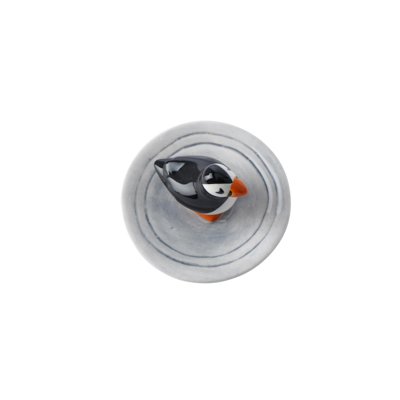 May include: A ceramic puffin figurine with a black and white body, orange beak, and white eye markings. The bird is perched on a round, gray, textured base with concentric circle patterns. The figurine is a decorative item.