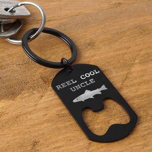 May include: Black metal bottle opener keychain with the text "REEL COOL UNCLE" and a fish graphic. The keychain is attached to a silver key ring and keys. The background is a wooden surface.