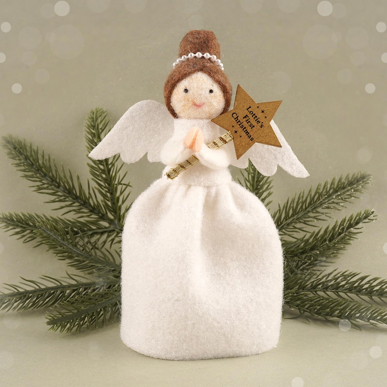 May include: A handcrafted Christmas angel figurine. The angel has a white dress, wings, and a brown hair bun adorned with a pearl headband. It holds a star-shaped sign that reads "Lottie's First Christmas". The background features a green pine branch.