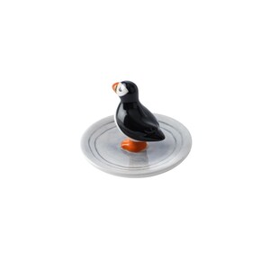 May include: A ceramic puffin figurine with a black body, white face, and orange beak and feet. The bird stands on a small, round, gray plate with subtle concentric circle detailing. The figurine is a decorative item.