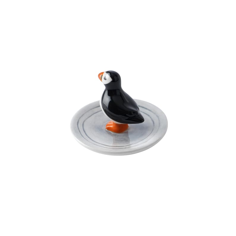 May include: A ceramic puffin figurine with a black body, white face, and orange beak and feet. The bird stands on a small, round, gray plate with subtle concentric circle detailing. The figurine is a decorative item.