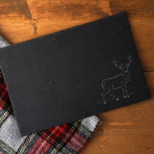 May include: A black slate serving board with a white outline of a deer on it. The board is lying on top of a red and black plaid fabric.