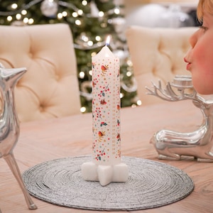 May include: A white advent candle with a star-shaped base. The candle is decorated with a festive pattern of red robins, holly berries, and gold stars. The numbers 1 through 24 are printed on the candle.
