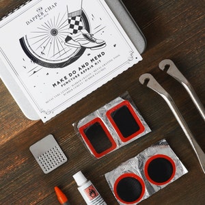 May include: A bicycle repair kit with a black and white illustration of a bicycle wheel and a shoe on the box. The kit includes metal tire levers, a marker crayon, a metal grating tool, rubber cement, and rubber patches. The kit is packaged in a white box with black text.