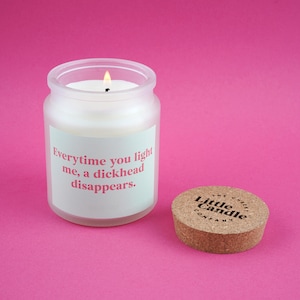 Pode incluir: Uma vela branca acesa em um frasco de vidro fosco com o texto "Everytime you light me, a dickhead disappears." Uma tampa de cortiça com "The Cheeky Little Candle Company" está ao lado da vela. O fundo é rosa.