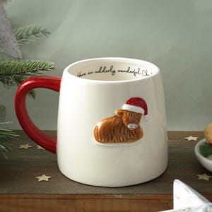 May include: White ceramic mug with a red handle. The mug features a brown highland cow wearing a red Santa hat on the front. The text "Have an utterly wonderful Christmas" is printed on the mug.