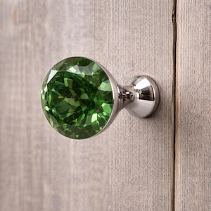 Può includere: Un pomello rotondo per mobili con una parte superiore in cristallo verde smeraldo sfaccettato, inserito in una base in metallo color argento lucido. Il cristallo circolare ha una superficie sfaccettata che riflette la luce.