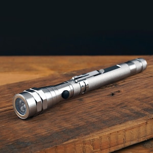 Könnte beinhalten: Eine silberne Taschenlampe mit strukturiertem Griff und hellem LED-Licht. Die Taschenlampe besteht aus Metall und hat einen schwarzen Knopf und einen Clip. Sie liegt auf einer Holzoberfläche.