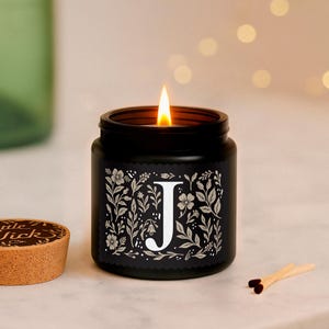 Puede incluir: Una vela negra encendida en un tarro con un dise&ntilde;o floral blanco y la letra "J". La vela est&aacute; sobre una superficie de m&aacute;rmol, con una tapa de corcho y cerillas. La llama de la vela es brillante.