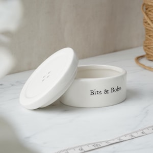 May include: White ceramic container with a lid shaped like a button. The container has the text "Bits & Bobs" printed on the side.