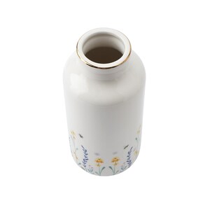 May include: White ceramic vase with a gold rim and a floral pattern featuring yellow flowers, green leaves, and blue stems. The vase has a narrow cylindrical shape.