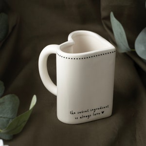 May include: White ceramic heart-shaped mug with a black handle. The mug has a black dotted line around the top and the text "the secret ingredient is always love" with a small heart at the end.