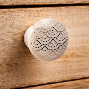 May include: A round white ceramic cabinet knob featuring a repeating gold dotted pattern of overlapping semicircles. The knob has a smooth, glossy finish and is attached to a light-colored wooden surface with a visible horizontal wood grain texture.
