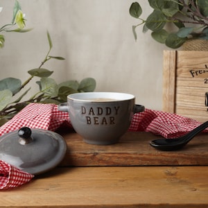 May include: A grey ceramic bowl with two handles and the text "Daddy Bear" on the front. The bowl is sitting on a wooden table with a red and white checked cloth and a black spoon.