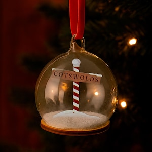 May include: A clear glass Christmas ornament with a red ribbon. Inside, a signpost reads "Cotswolds" above a red and white striped pole, set in a snowy base. The ornament is round and has a gold-colored base.