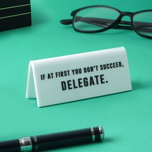 May include: White desk sign with black text that reads "If at first you don't succeed, delegate."