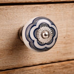 May include: A round ceramic cabinet knob featuring a white base with a dark blue and grey floral pattern. The center of the knob is adorned with a silver-toned metallic circular plate with a ridged edge and a smooth, raised silver-toned dome.